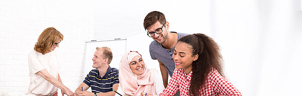 junge Erwachsene unterschiedlicher Kulturellen sitzen an einem Tisch, eine Lehrerin und ein Lehrer schauen ihnen über die Schulter und helfen bei Aufgaben